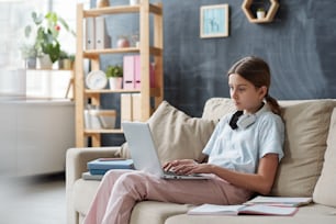 Adolescente serio con computadora portátil sentado en el sofá en el entorno doméstico y navegando en la red mientras estudia de forma remota durante la cuarentena