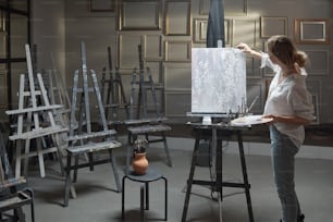 Junge blonde Frau in Freizeitkleidung, die die Farbpalette hält, während sie vor dem Bild auf der Staffelei steht und im Atelier malt