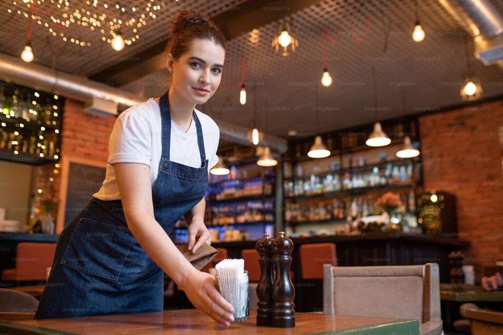 Hübsche junge Kellnerin in Arbeitskleidung, die sich über den Tisch beugt, während sie Glas mit Zahnstochern aufstellt und Sie im Café oder Restaurant ansieht