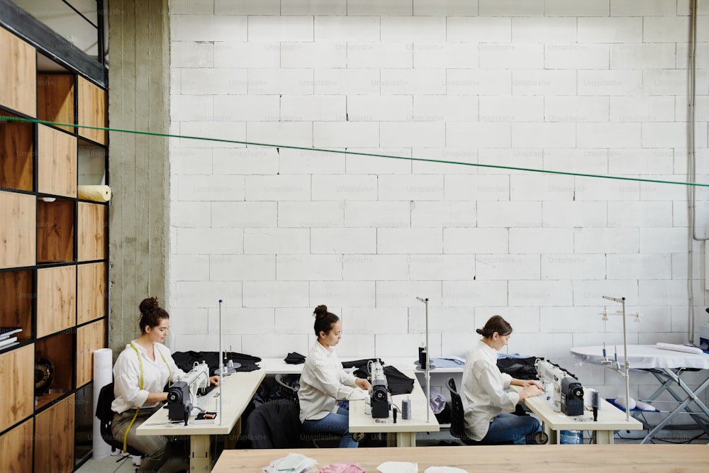 세 명의 젊은 현대 여성 디자이너가 작업장에서 작업장에 앉아 있는 동안 새로운 sesonal 컬렉션을 위한 패션 재봉 아이템