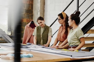 Two young trainees of tailor showing her new sketches of fashion items and consulting with her about details at start-up meeting