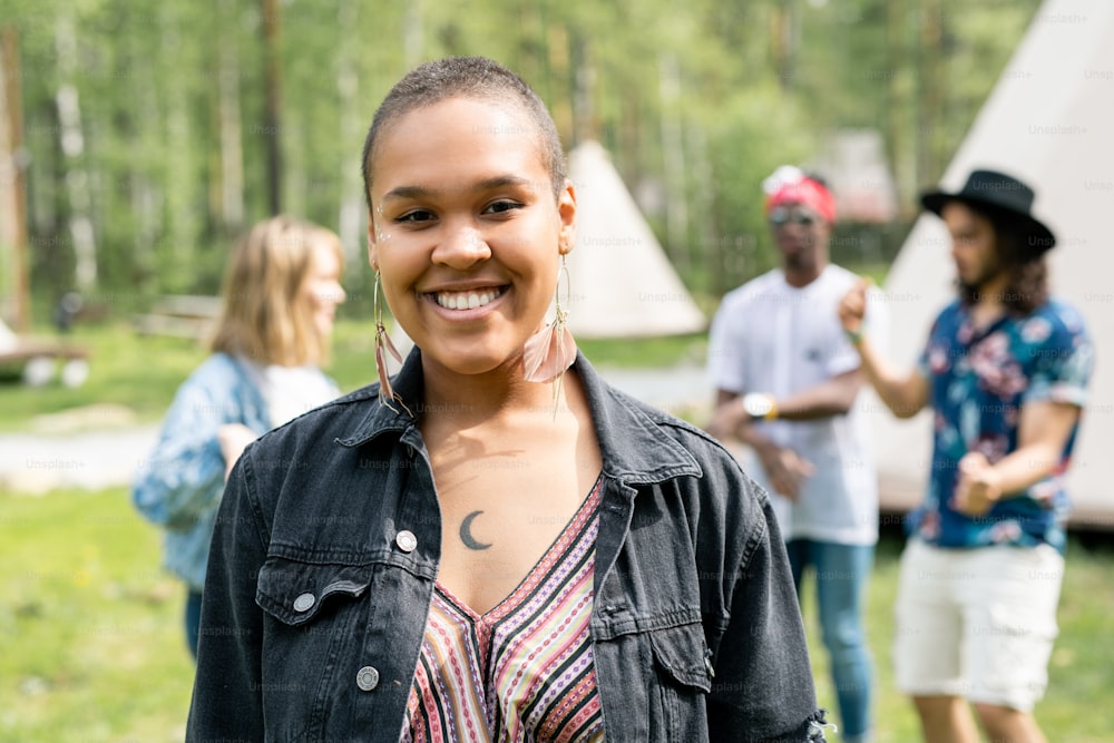 Ritratto di ragazza afroamericana sorridente in orecchini di pelle in piedi contro gli amici al campeggio