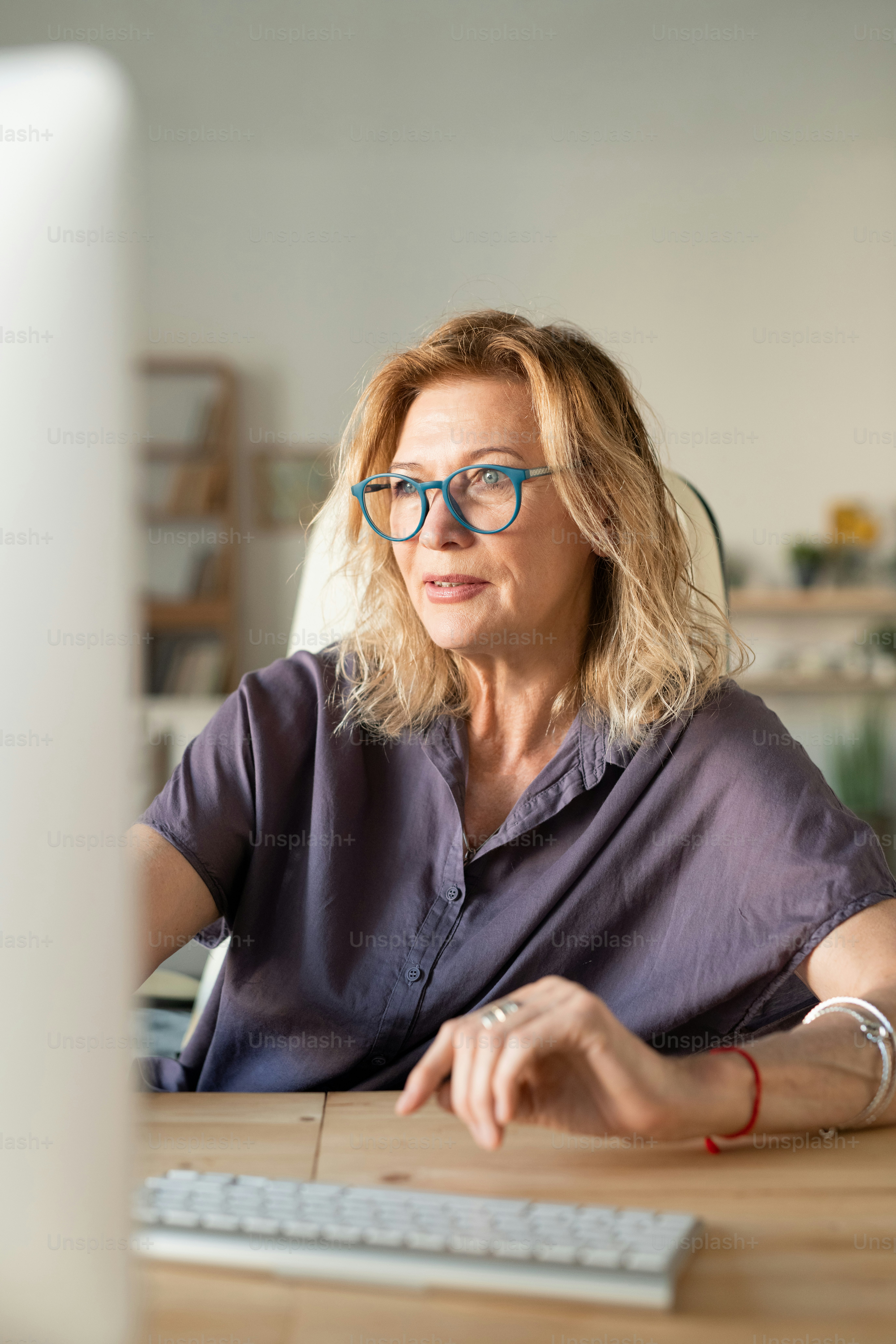 Empresaria rubia madura en ropa casual mirando la pantalla de la computadora mientras está sentada junto a la mesa frente al monitor y trabajando en casa