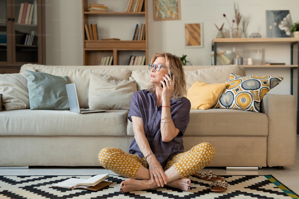 Donna matura in abbigliamento casual seduta sul tappeto vicino al divano con le gambe incrociate mentre parla con il suo amico o marito sullo smartphone