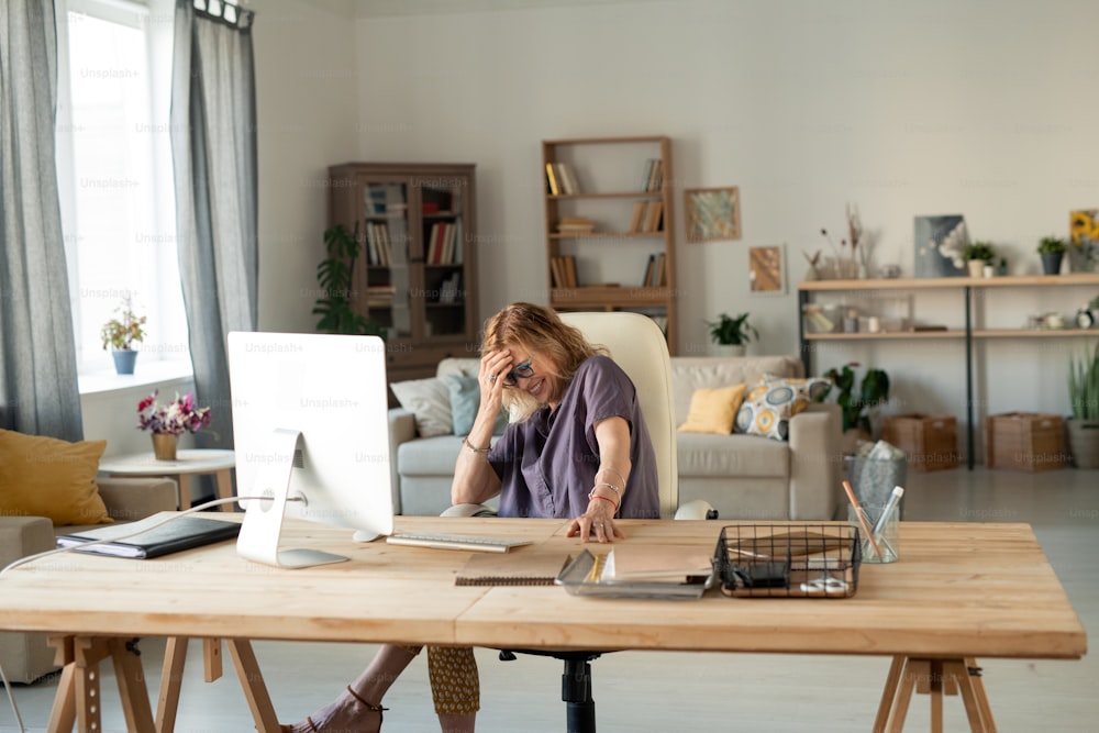 Fêmea loira madura e alegre rindo na frente da tela do computador enquanto se senta à mesa na frente do monitor e assiste a filmes on-line em casa