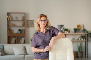Contemporary mature businesswoman in eyeglasses standing by white leather armchair by her workplace in front of camera at home