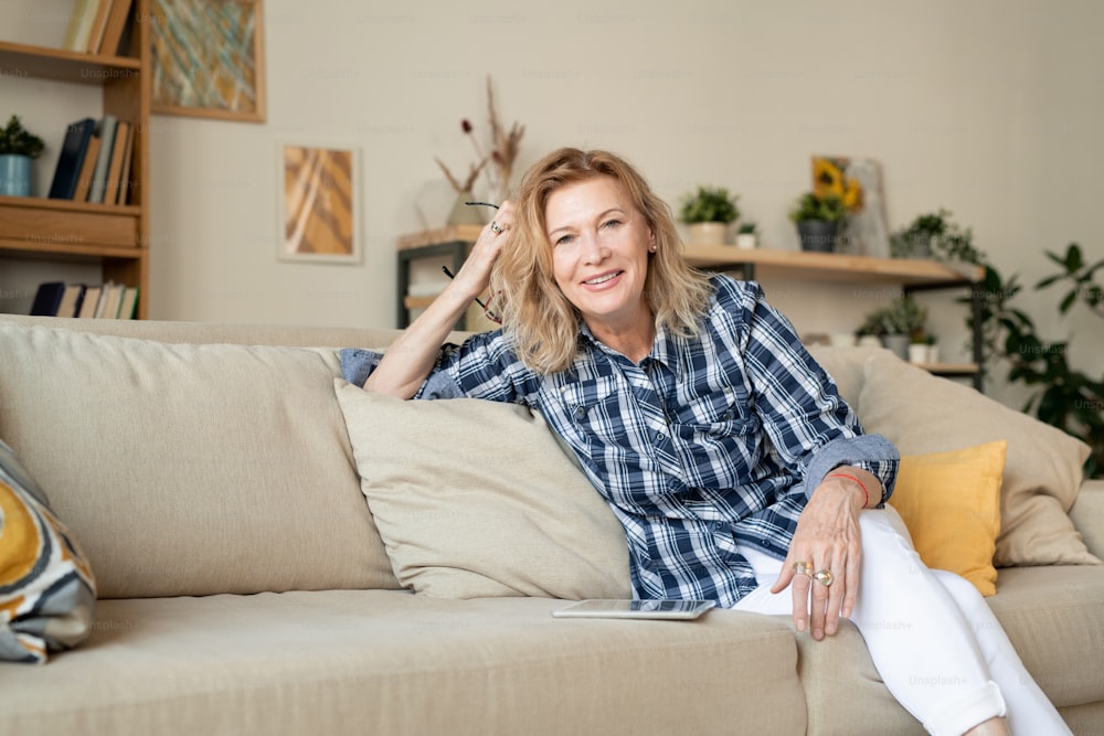 Alegre mujer madura rubia con jeans blancos y camisa a cuadros sentada en un cómodo sofá frente a la cámara en el entorno del hogar