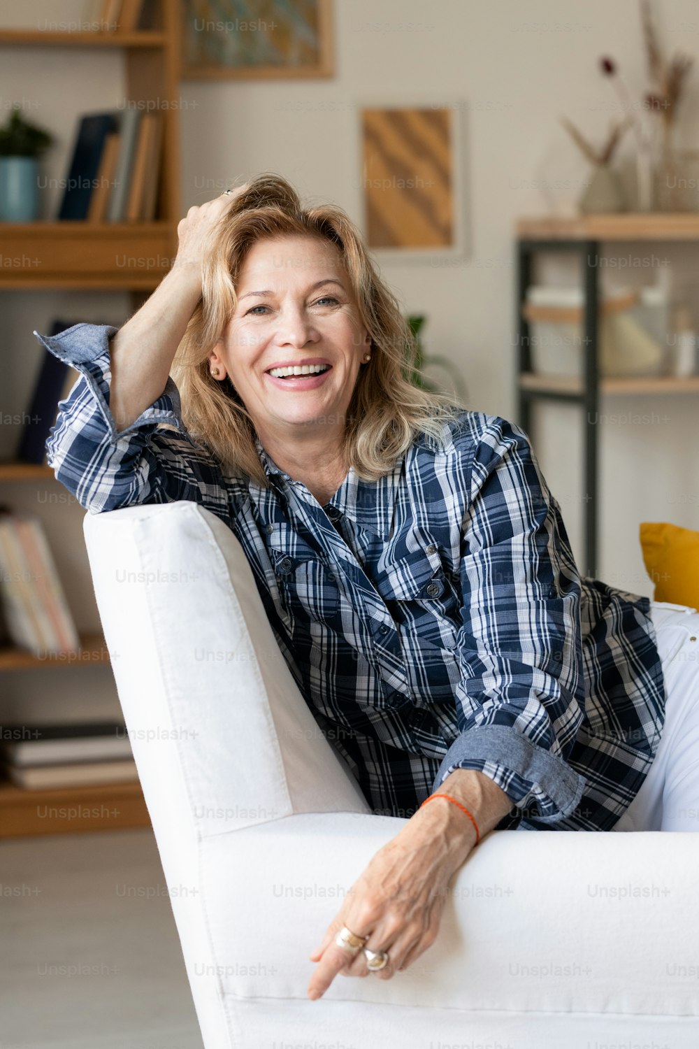 Reife lachende Frau in Freizeitkleidung, die dich mit einem zahnigen Lächeln ansieht, während sie in einem weißen Sessel vor der Kamera im Wohnzimmer sitzt