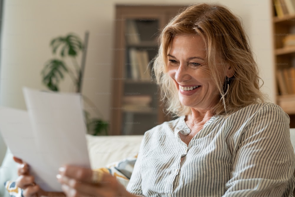 Mujer madura feliz riendo mientras mira fotos familiares mientras se queda en casa el fin de semana o se relaja por la noche después del día de trabajo