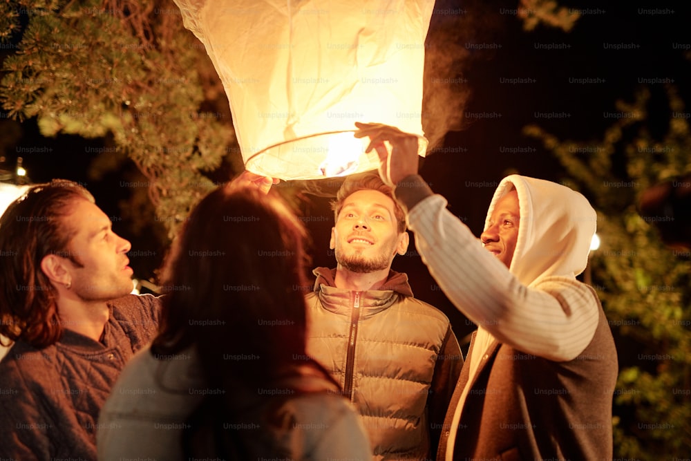 친절한 젊은 다문화 남성과 여성은 자연 환경에서 밤에 조명이 켜진 큰 흰색 풍선을 바라보고 있다
