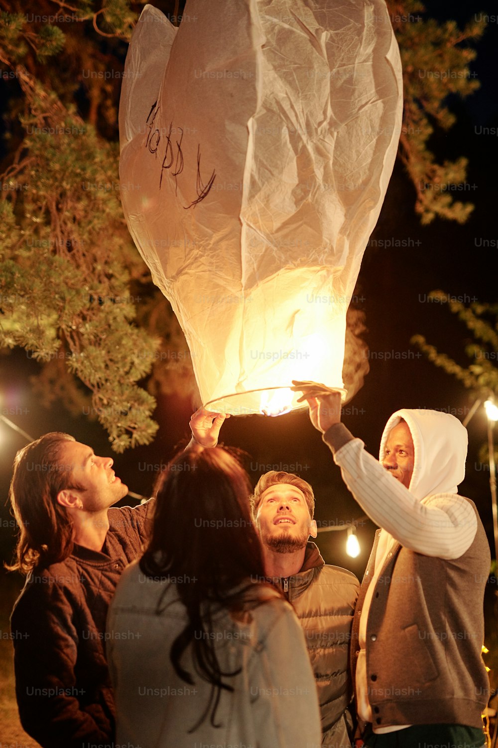 캐주얼웨어를 입은 다문화 젊은 친구들이 밤에 소나무 아래에서 모여 조명이 켜진 대형 풍선을 들여다보고 있다
