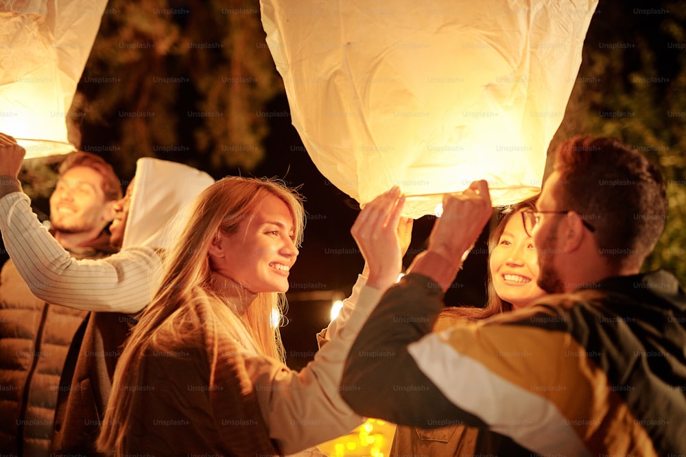 Giovani amici sorridenti interculturali che tengono grandi palloncini bianchi con illuminazione mentre si godono la festa notturna in un ambiente naturale