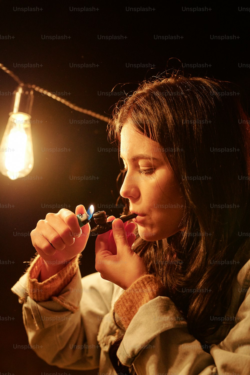 Young brunette female lighting tobacco pipe while holding lighter over its wide part and smoking against burning lamp outdoors