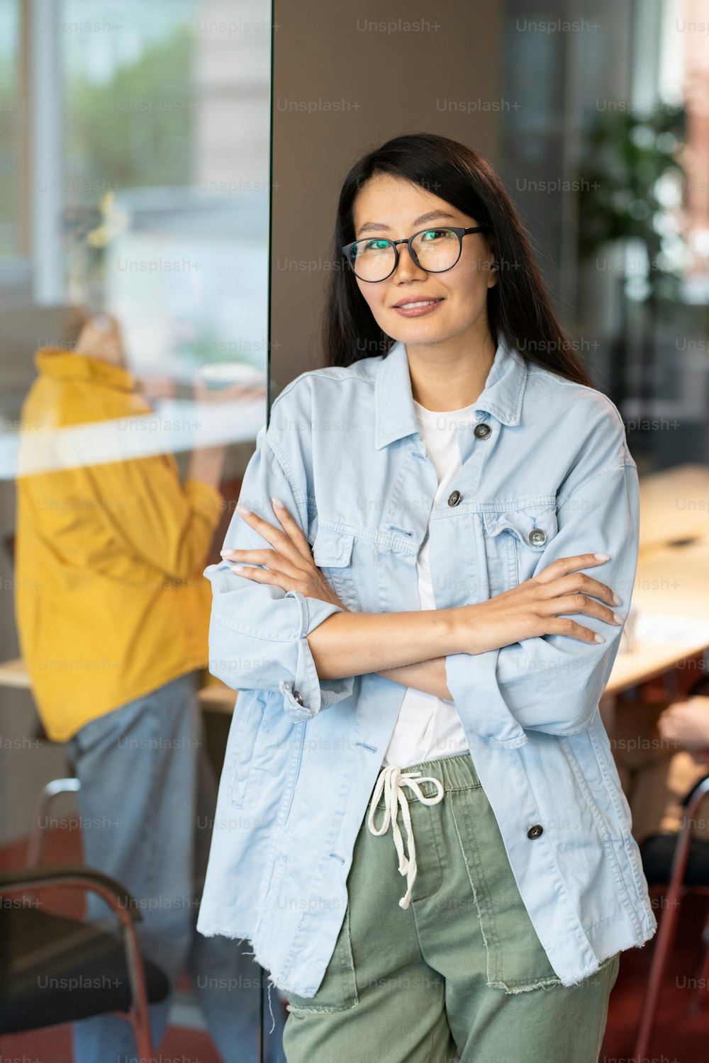 Happy young Asian businesswoman crossing her arms by chest while standing in front of camera against colleague behind office wall
