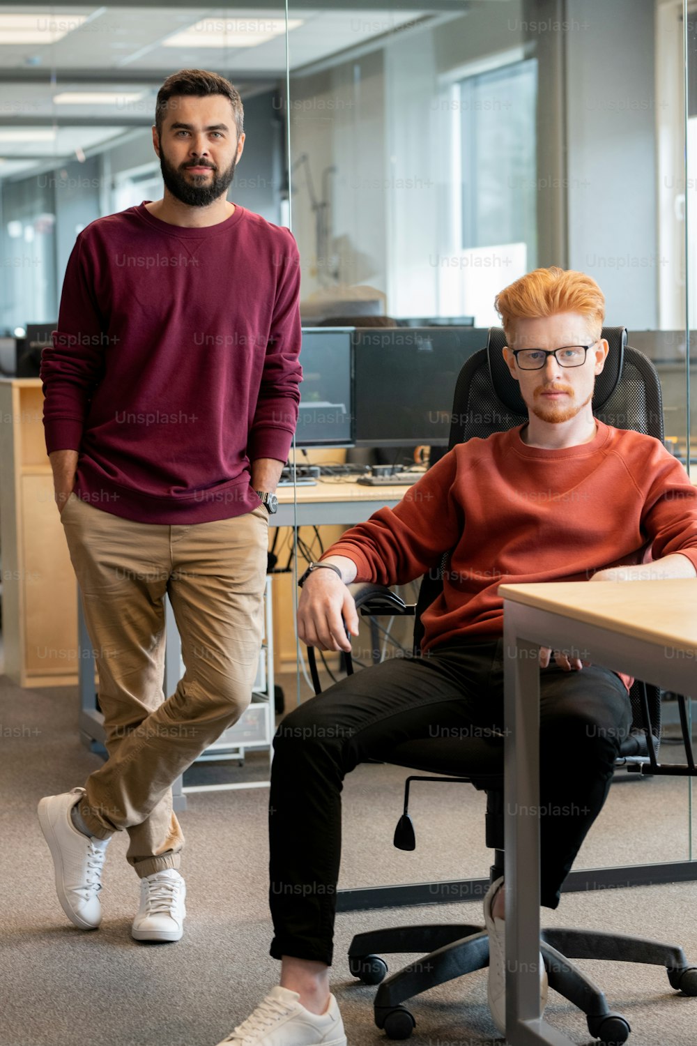 Two young businessmen in casualwear sitting and standing by workplace in front of camera against monitors in large contemporary office