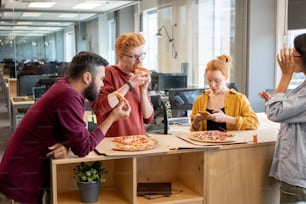 Group of young colleagues in casualwear talking and using smartphone at lunch break while having pizza by table in large contemoprary office