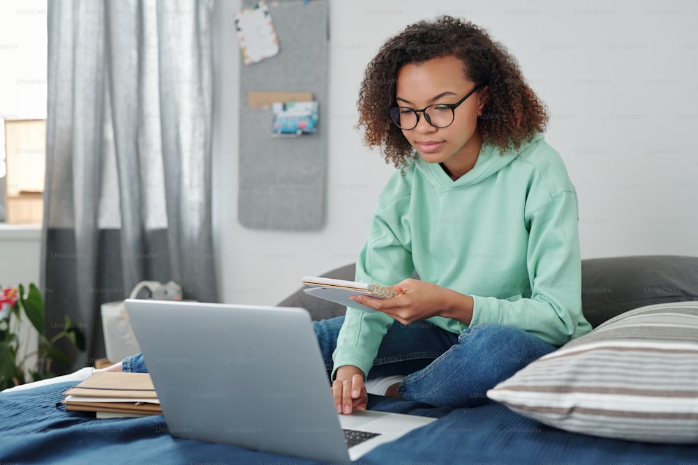 Jovem estudante mestiça em trajes casuais sentada na cama em frente ao laptop, lendo anotações em copybook e navegando na rede em casa