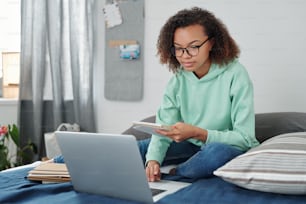 Young mixed-race female student in casualwear sitting on bed in front of laptop, reading notes in copybook and surfing in the net at home