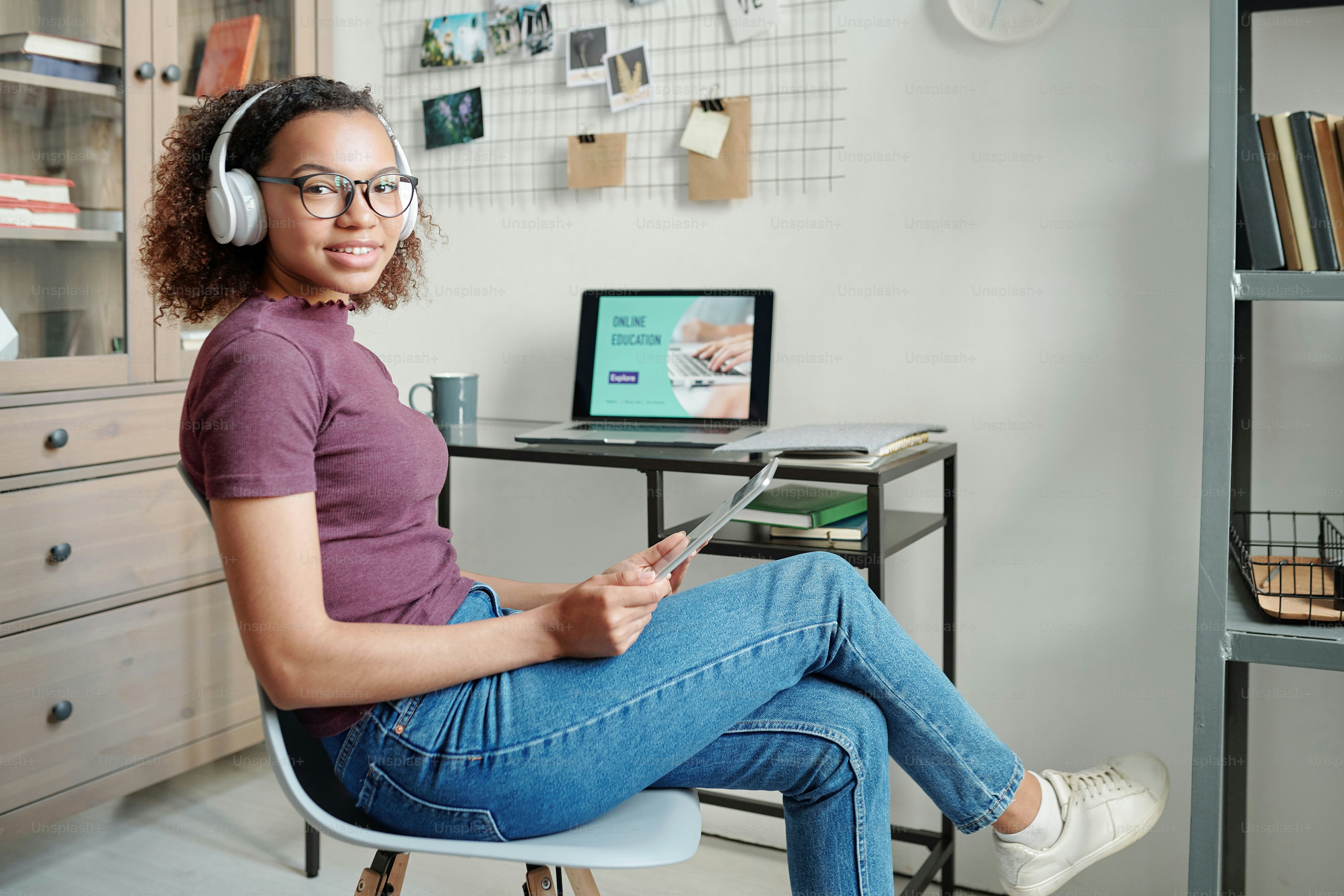 Wanita ras campuran muda yang ceria dengan pakaian kasual dan headphone menatap Anda di depan laptop saat menggunakan touchpad di lingkungan rumah