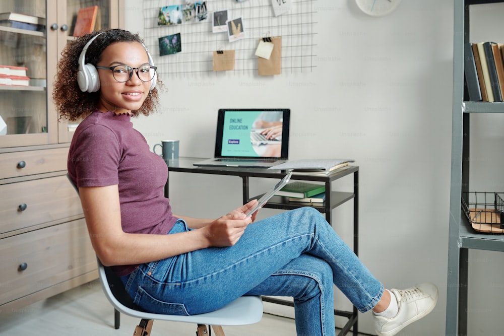Jeune femme métisse joyeuse en tenue décontractée et écouteurs vous regardant contre un ordinateur portable tout en utilisant le pavé tactile dans un environnement domestique