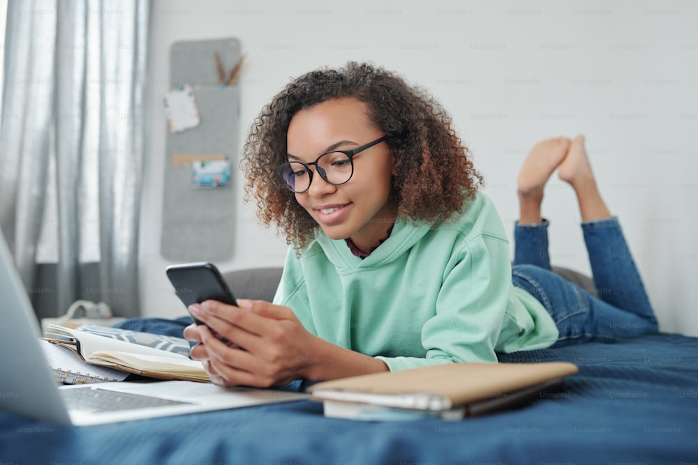 Young smiling mixed-race student in jeans and hoodie scrolling in smartphone while lying on bed in front of laptop in her bedroom