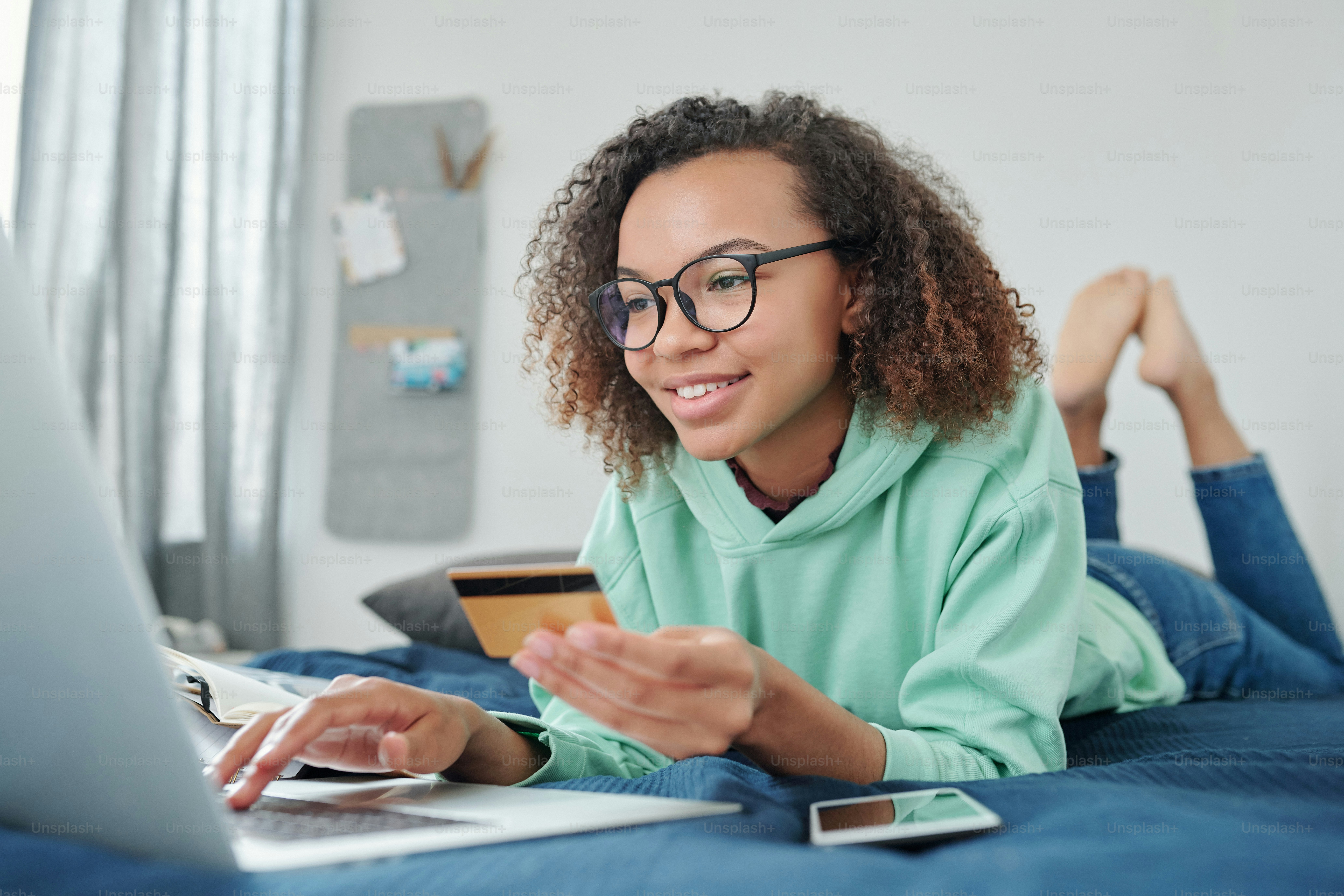 Wanita ras campuran muda yang bahagia dengan kacamata dan pakaian kasual berbaring di tempat tidur di depan laptop dan menggulir barang-barang di toko online