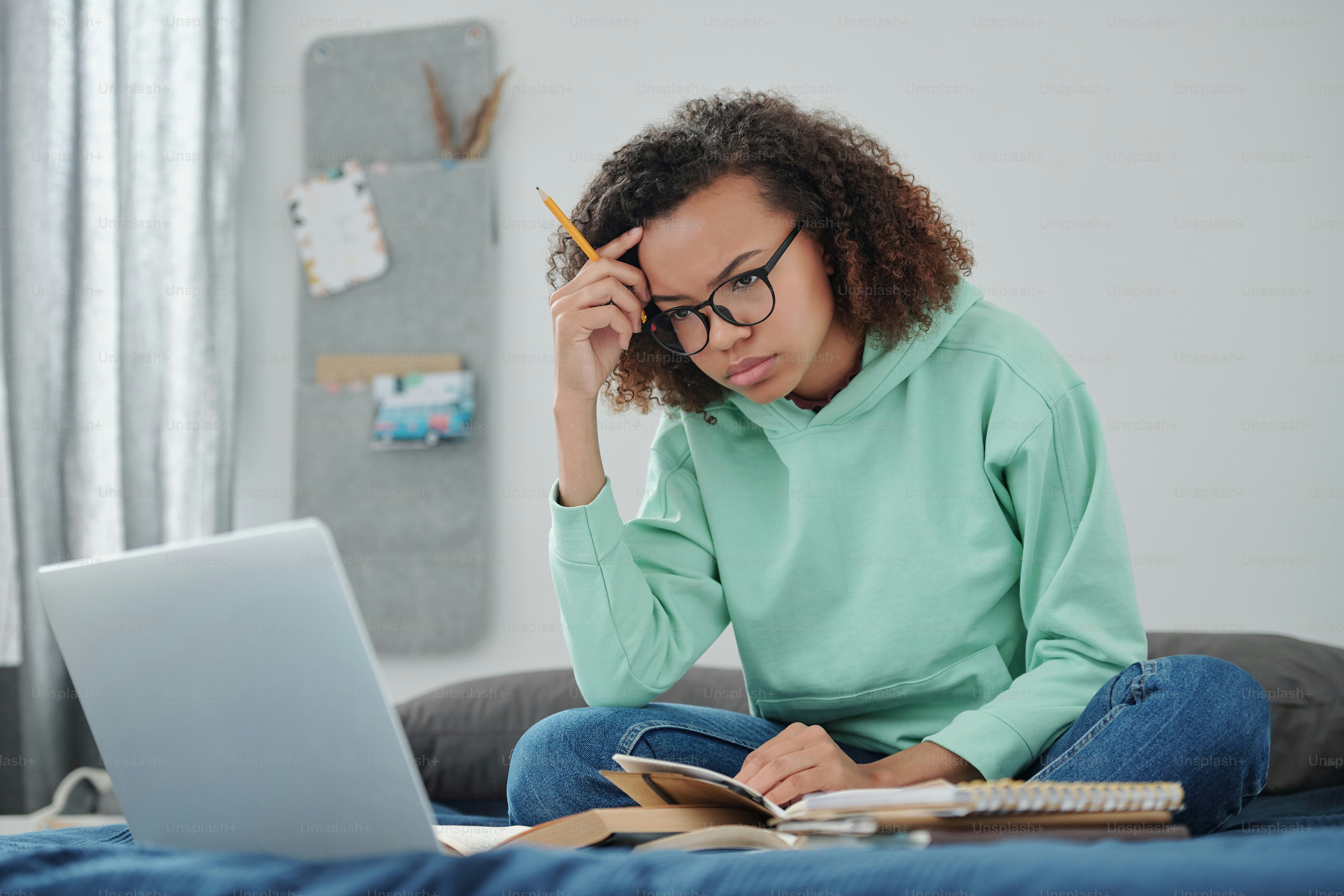 Mahasiswi muda dengan celana jeans dan hoodie mencoba berkonsentrasi sambil duduk di tempat tidur di depan laptop dan mempersiapkan ujian yang akan datang