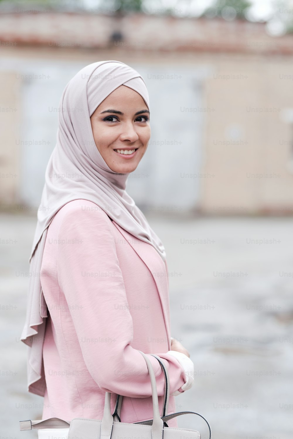 히잡과 핑크색 가디건을 입은 젊은 쾌활한 무슬림 여성이 핸드백을 들고 도시 환경에서 카메라 앞에서 웃고 있다