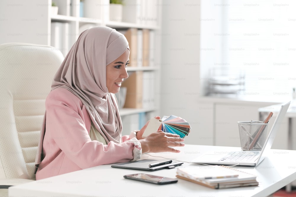 Joven diseñadora musulmana con hijab y ropa casual inteligente que muestra la paleta a uno de los clientes en la pantalla de la computadora portátil durante el trabajo en la oficina