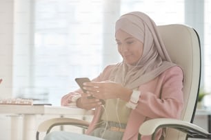 Young smiling Muslim businesswoman in hijab scrolling in smartphone while sitting in armchair and looking for contacts or messaging