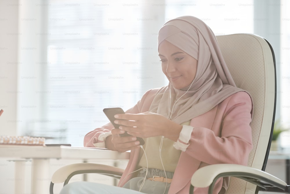 Joven mujer de negocios musulmana sonriente con hijab desplazándose en un teléfono inteligente mientras está sentada en el sillón y busca contactos o mensajes