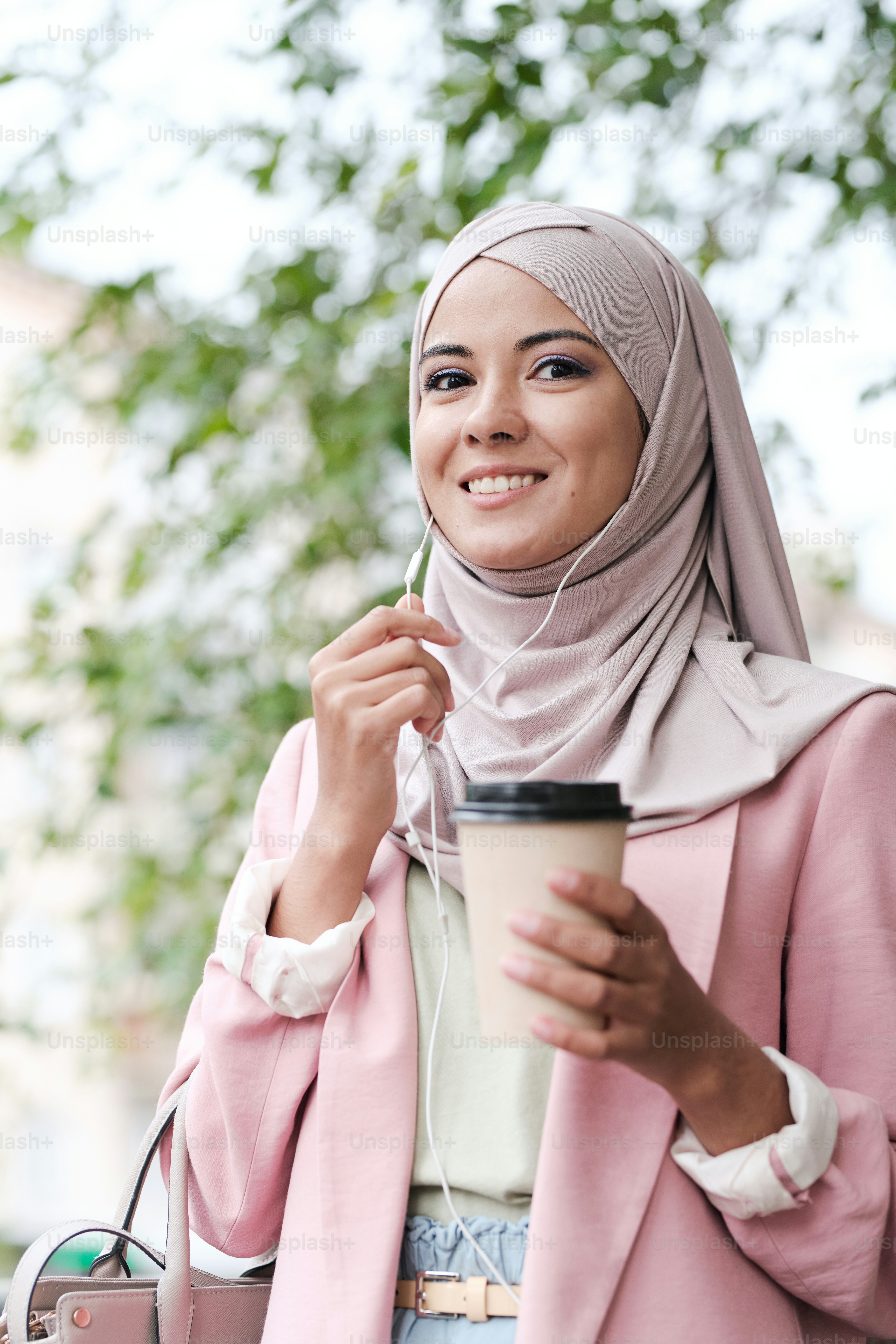Wanita muda cantik tersenyum Muslim dengan segelas kopi berbicara dengan seseorang di telepon sambil berdiri di lingkungan perkotaan