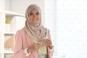 Feliz joven empresaria con hijab y ropa casual elegante sosteniendo un lápiz óptico mientras está parada frente a la cámara en la oficina y te mira con una sonrisa