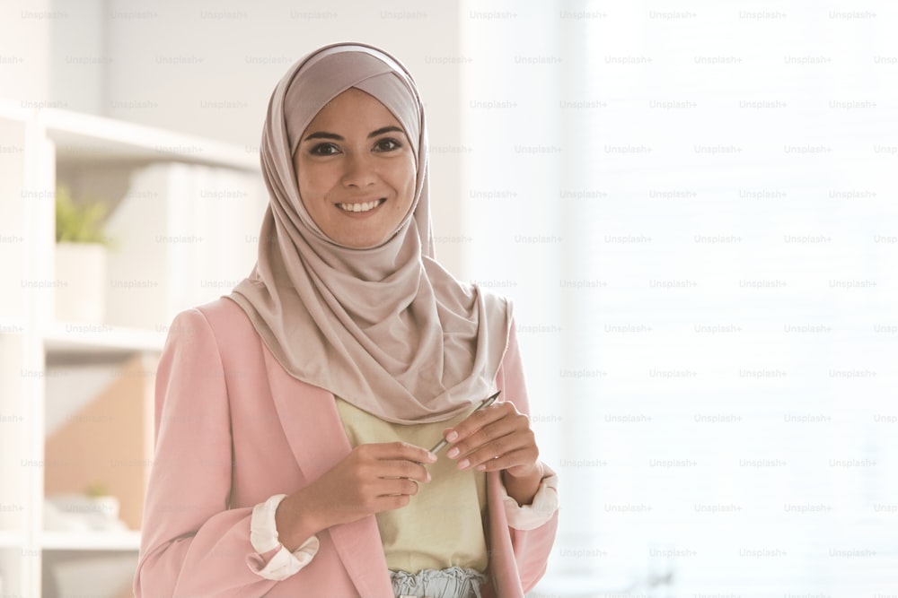 Giovane donna d'affari felice in hijab e abbigliamento casual elegante che tiene lo stilo mentre si trova davanti alla telecamera in ufficio e ti guarda con un sorriso