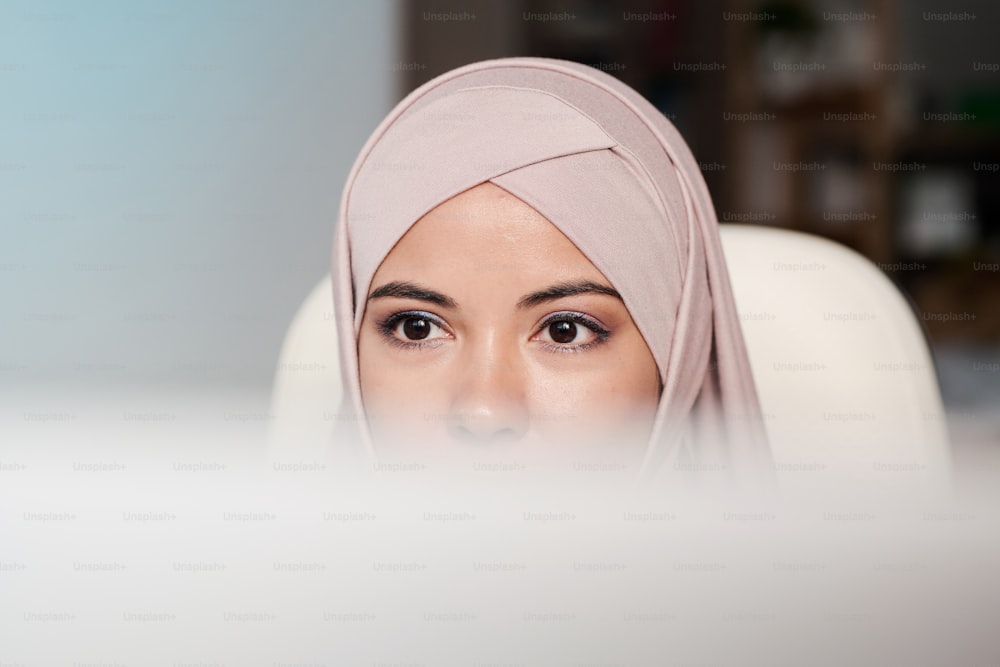 Parte superior da cabeça da jovem empresária contemporânea no hijab sentada na poltrona na frente do monitor do computador e olhando para a tela
