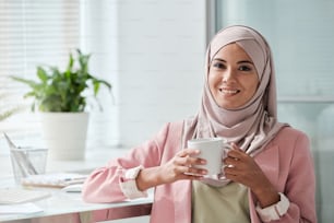 Junge erfolgreiche muslimische Geschäftsfrau in Hijab und eleganter Freizeitkleidung, die am Arbeitsplatz vor der Kamera steht und Tee oder Kaffee trinkt