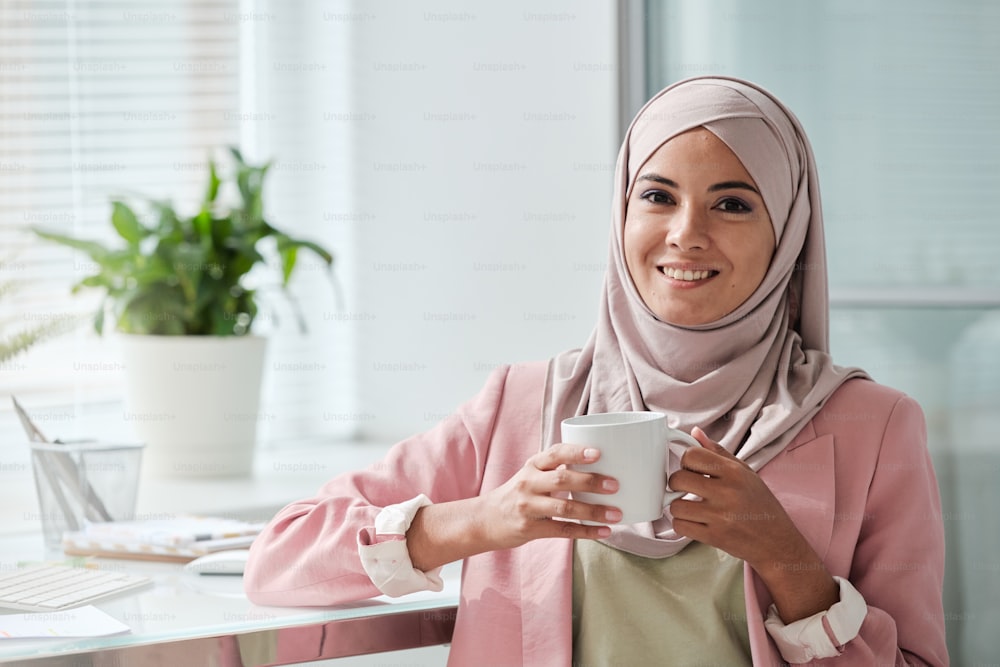 히잡을 쓰고 우아한 캐주얼웨어를 입은 젊은 무슬림 사업가가 카메라 앞에서 직장에 서서 차나 커피를 마시고 있다