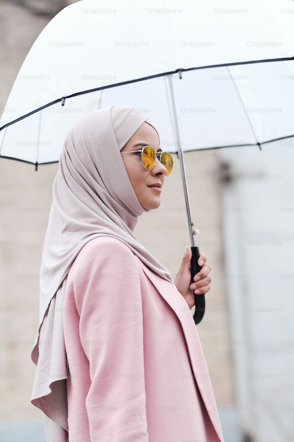Young stylish Muslim woman in hijab, sunglasses and pink cardigan holding umbrella while standing in the rain in urban environment