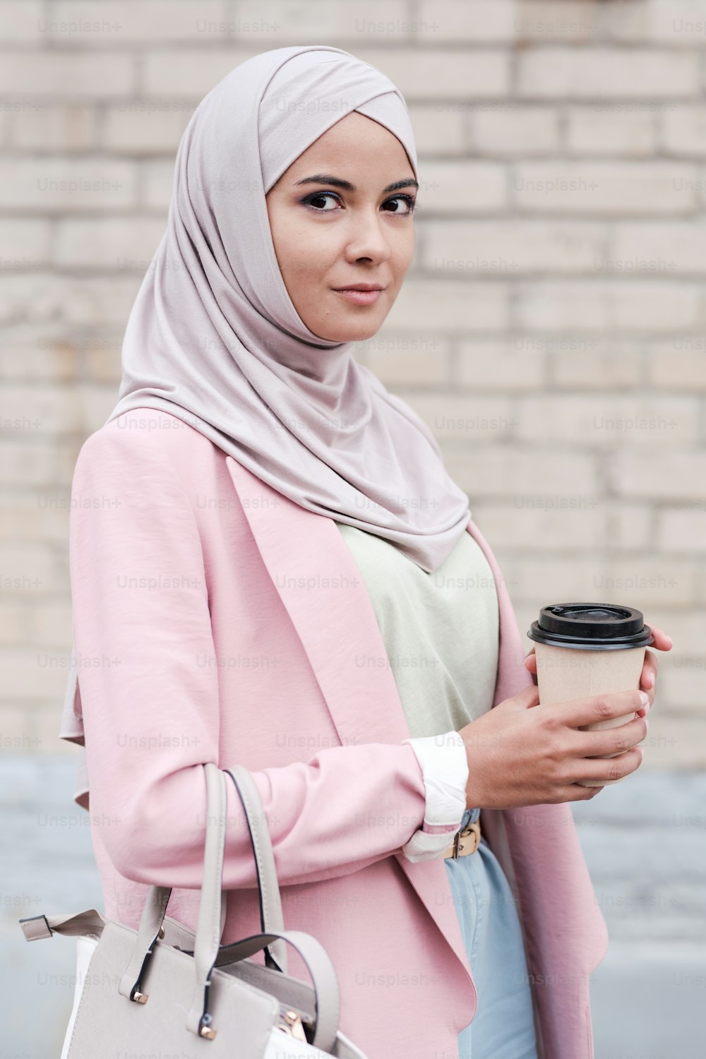 히잡, 풀오버, 핑크 가디건을 입은 젊은 무슬림 여성이 도시 환경에서 카메라 앞에 서서 커피를 마시고 있다