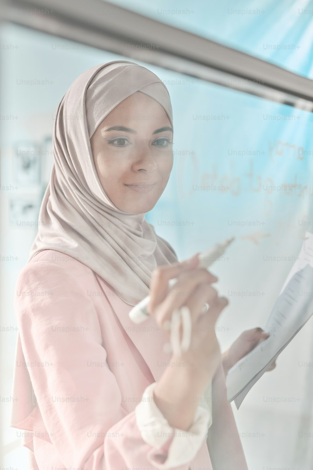 Joven corredora exitosa en hijab preparándose para la presentación o seminario mientras toma notas con resaltador en tablero transparente