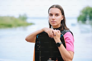 Active young female surfer in sportswear holding surfboard while standing against waterside in natural environment in front of camera