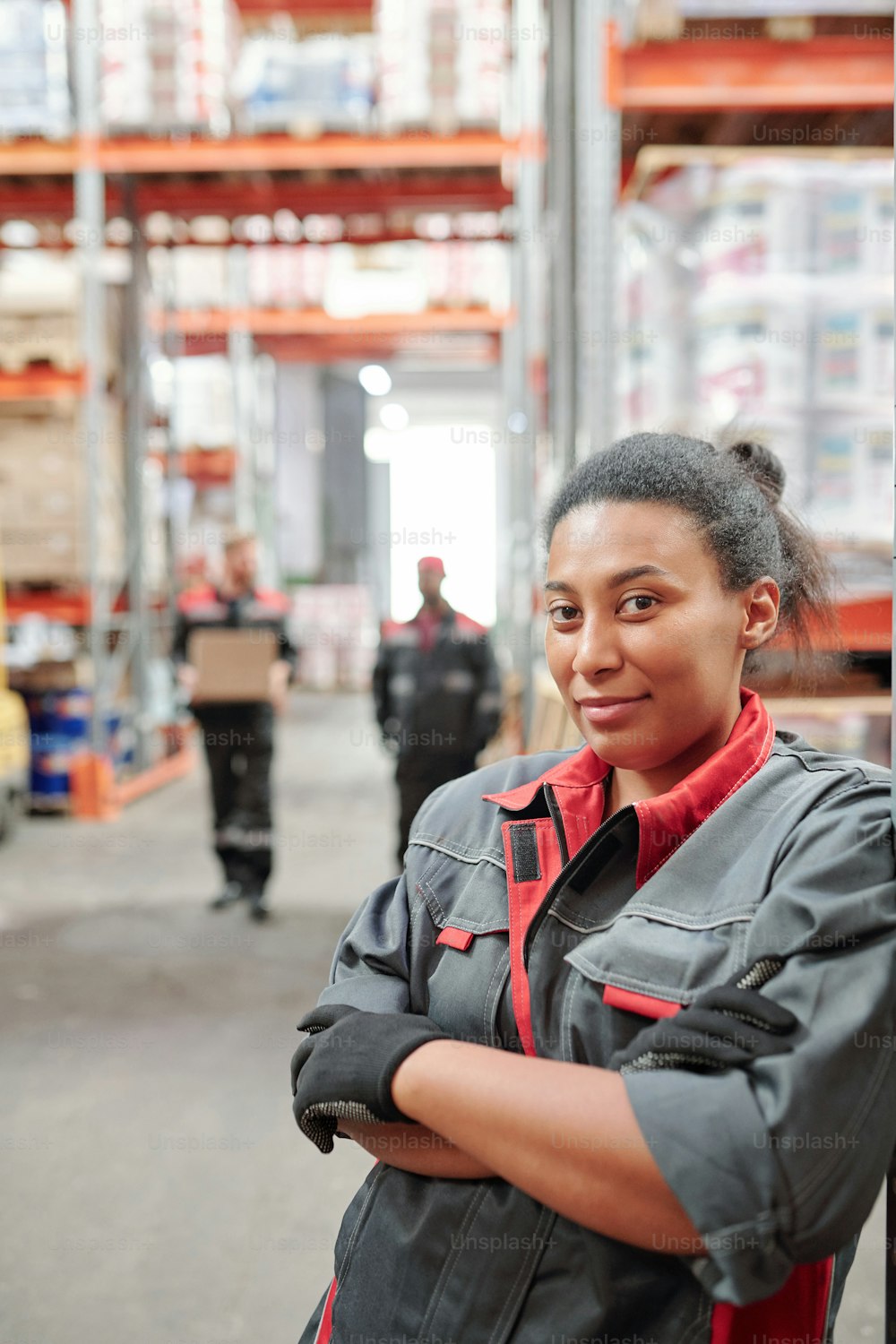 Successful young mixed-race female worker of large warehouse crossing arms by chest while standing by rack in aisle against male colleagues