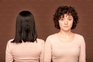 Young serious brunette woman of Caucasian ethnicity in white pullover standing in front of camera on background of back of another female