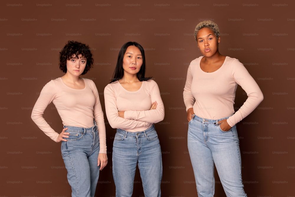 Trois jeunes femmes interculturelles en pulls blancs et jeans bleus debout en rangée sur fond brun et vous regardant