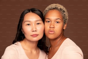Two young intercultural females looking at camera while standing close to one another and touching by their faces against brown background