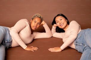 Due giovani donne contemporanee di varie etnie in blue jeans e pullover bianchi che ti guardano con sorrisi a denti stretti mentre sono sdraiate sul pavimento