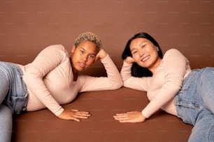 Two young contemporary females of various ethnicities in blue jeans and white pullovers looking at you with toothy smiles while lying on the floor