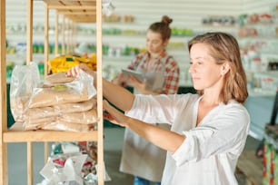 Jolie femme brune en tenue décontractée et lunettes debout près d’une étagère avec des engrais emballés et en choisissant un pour son jardin au supermarché