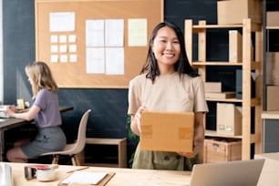 Young smiling female worker of online shop office with packed box looking at you while standing by workplace against colleague using laptop