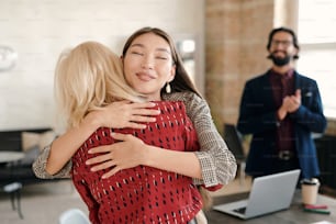 Happy Asian businesswoman congratulating one of colleagues on great work while embracing her against young elegant man applauding
